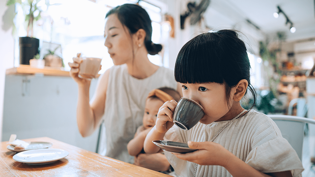 a little girl drinks a babycino at a cafe, she is sitting with her mom (also drinking coffee) who is holding a baby